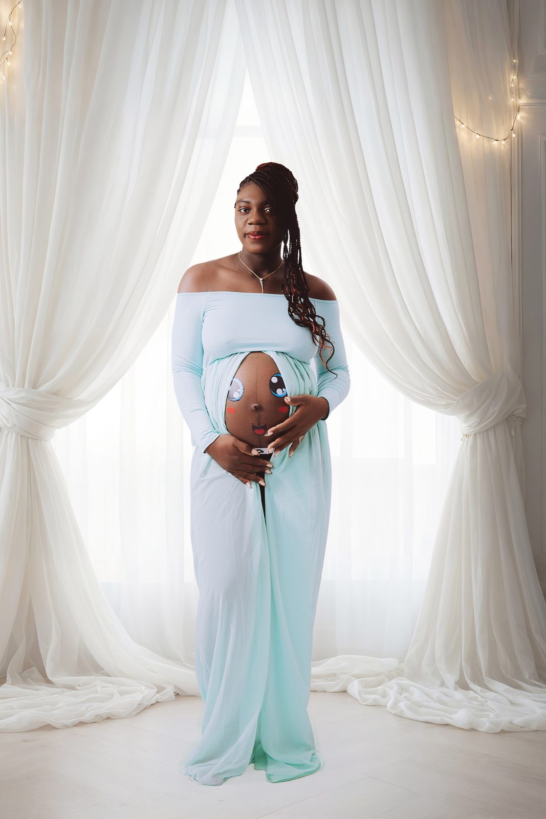 Future maman en robe bleu pastel, ventre peint d'un visage souriant, rideaux blancs